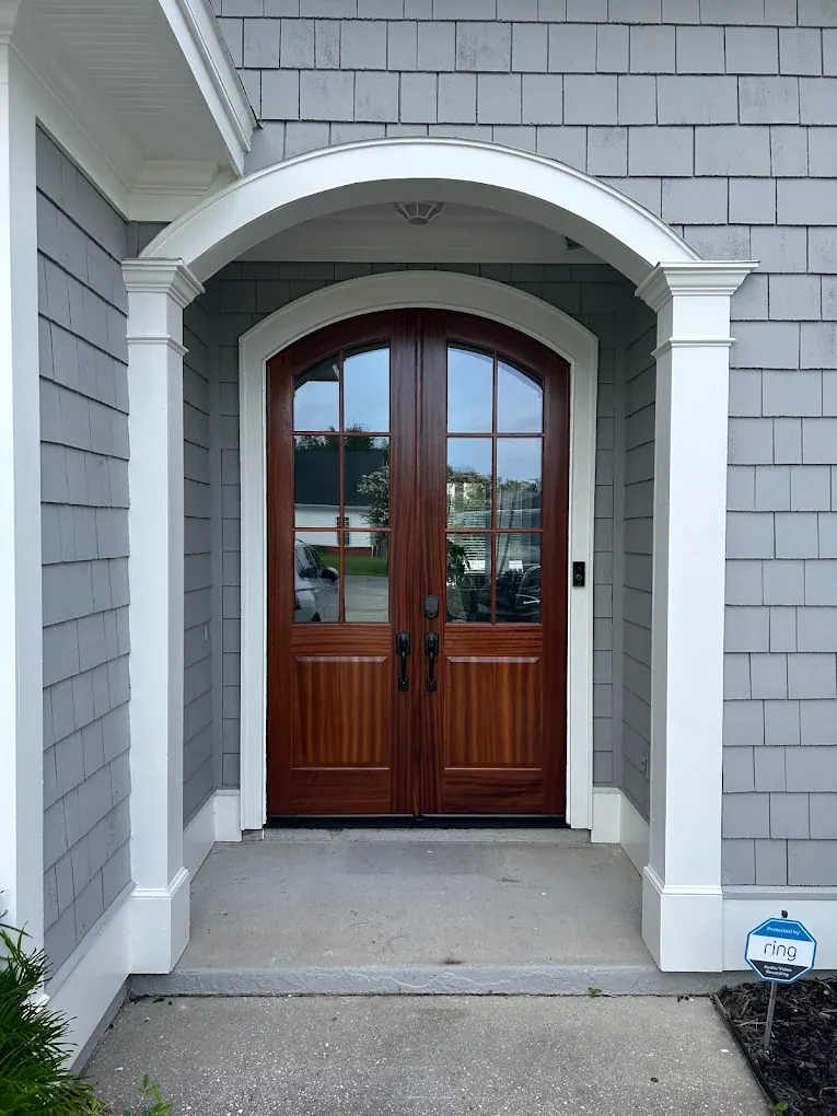 Arched wood double front doors with divided-lite glass and black handle sets, under a white columned arch on a gray shake-siding home.