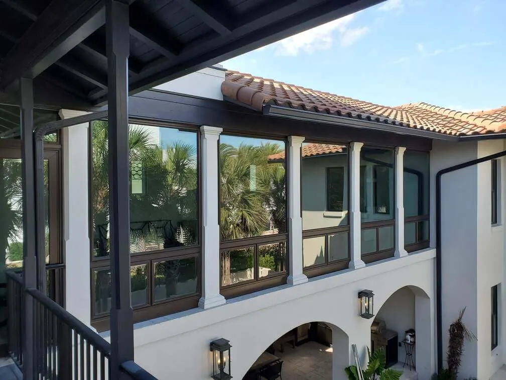 Second-floor loggia enclosed with dark bronze-framed windows between white columns on a stucco home with a clay tile roof.