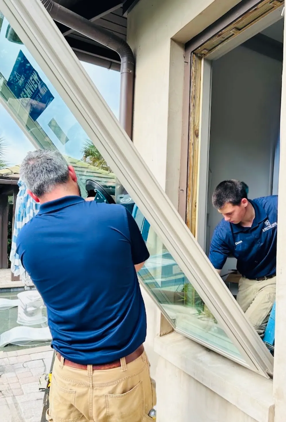 Two A Better View installers in navy company polos guiding a large window unit into a wall opening; the installer on the right wears a "Dean" name patch.
