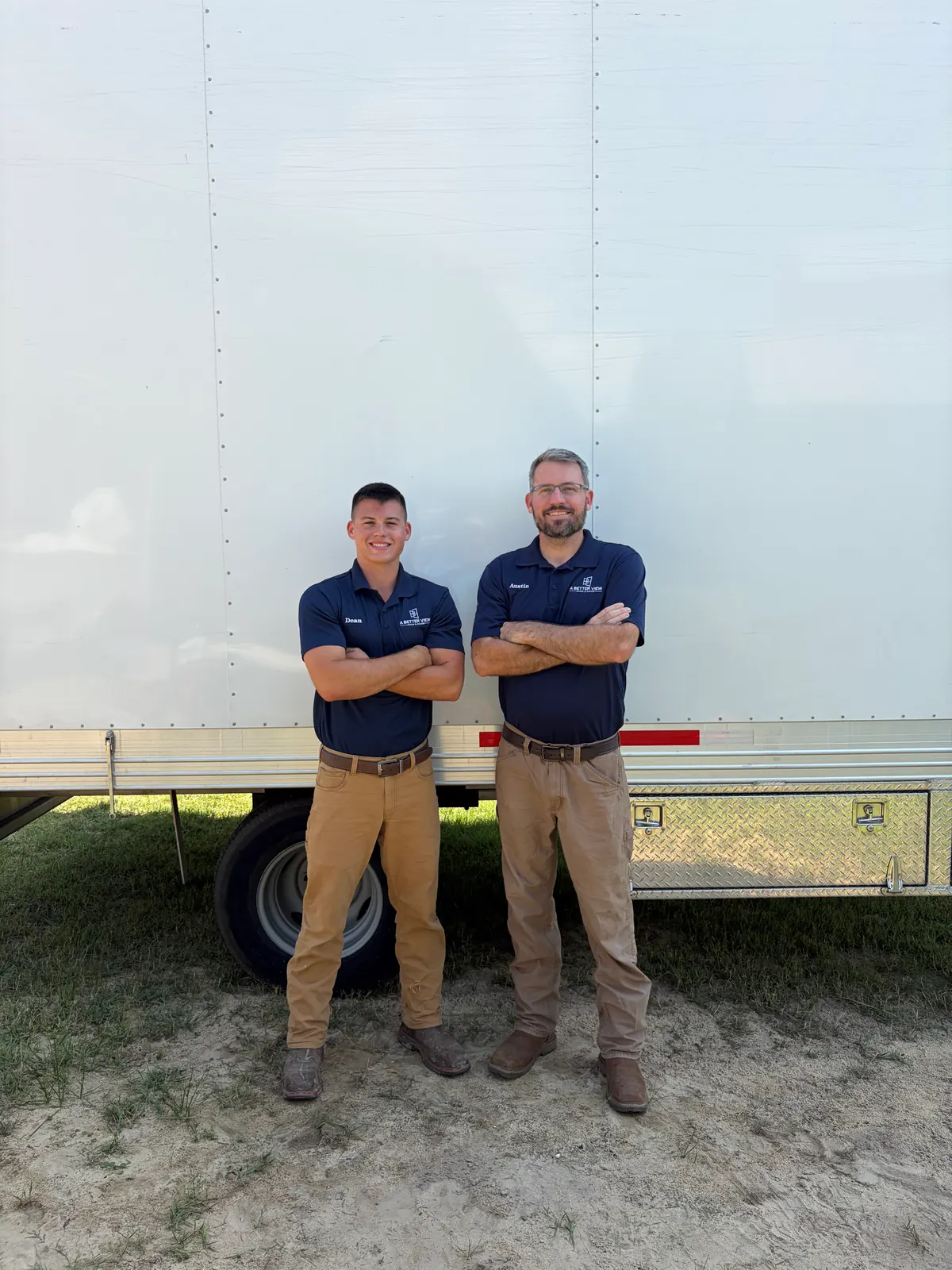 Dean Boyd and Austin Boyd standing with arms crossed in navy A Better View polos and khaki pants in front of a white box trailer.