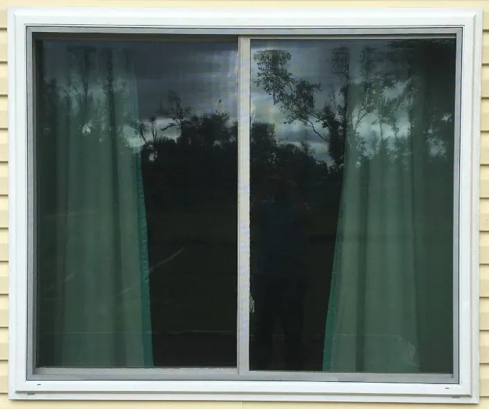 Two-panel horizontal sliding window with a white frame on pale yellow vinyl siding; the glass reflects trees and the photographer.