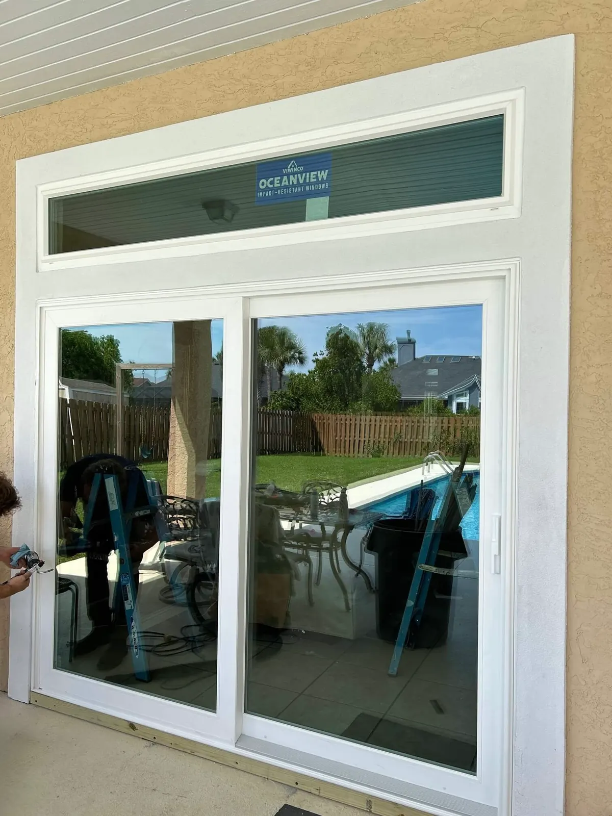 White sliding glass door with a transom window above, labeled Viwinco Oceanview impact-resistant, in a tan stucco wall facing a pool; an installer is caulking at the left edge.