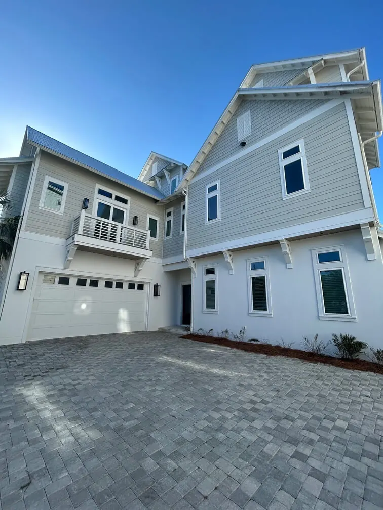 Newly built three-story coastal home with gray lap siding, white trim, dark-framed windows, a balcony and a paver driveway.