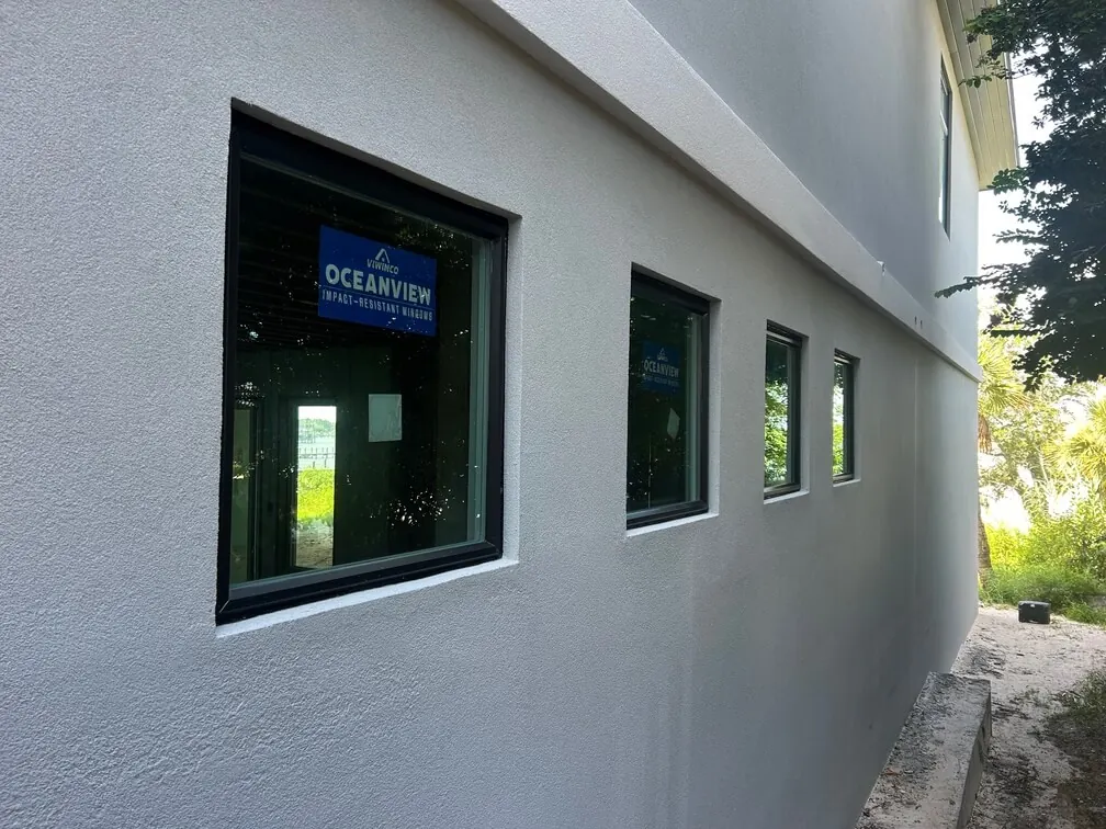 Row of black-framed windows with Viwinco Oceanview impact-resistant window labels on a freshly finished gray stucco wall.