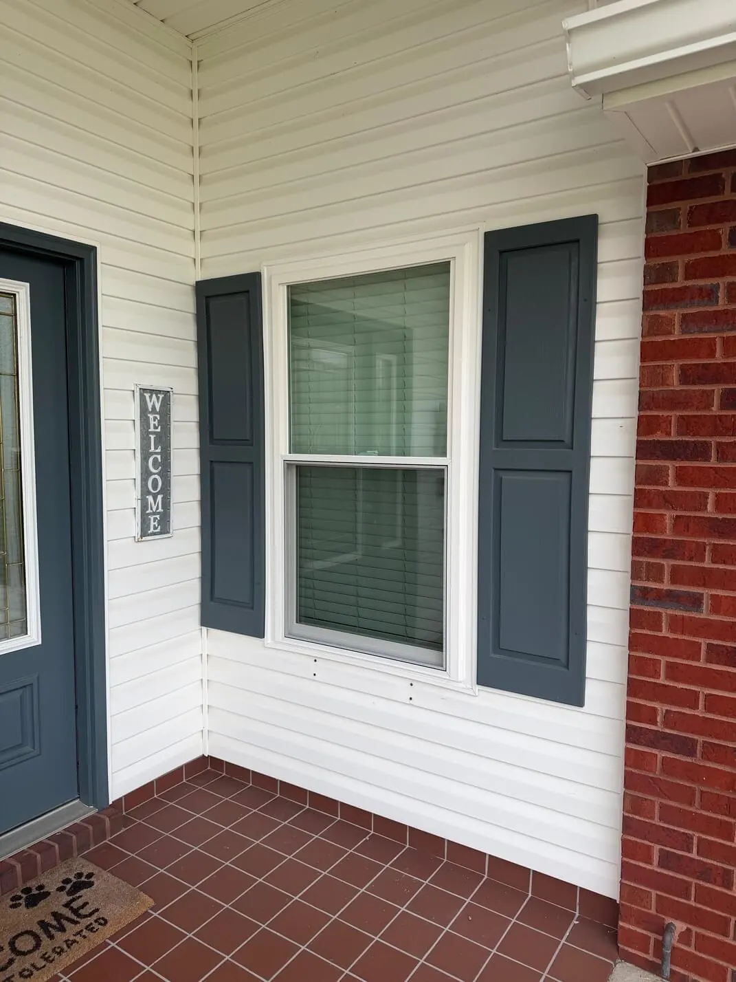 White double-hung window with dark gray shutters on white vinyl siding beside a front door and a red tile porch.