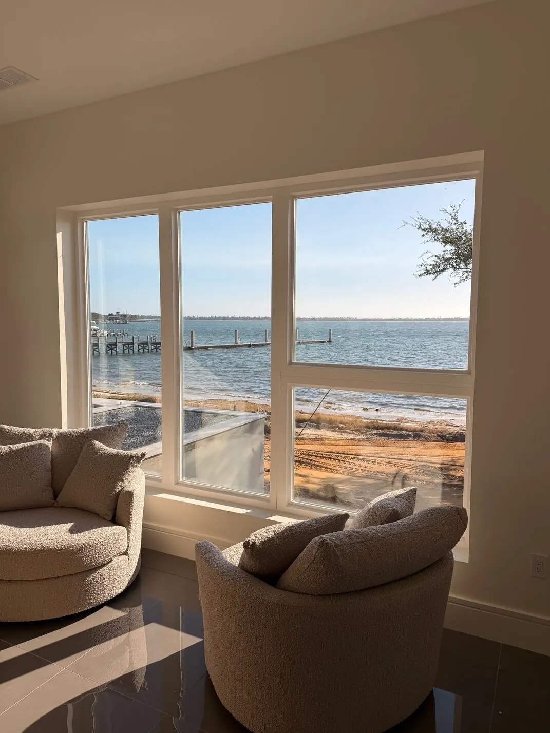 Large white-framed window combination in a sunlit living room looking out over a bay with wooden docks, two round chairs in front.